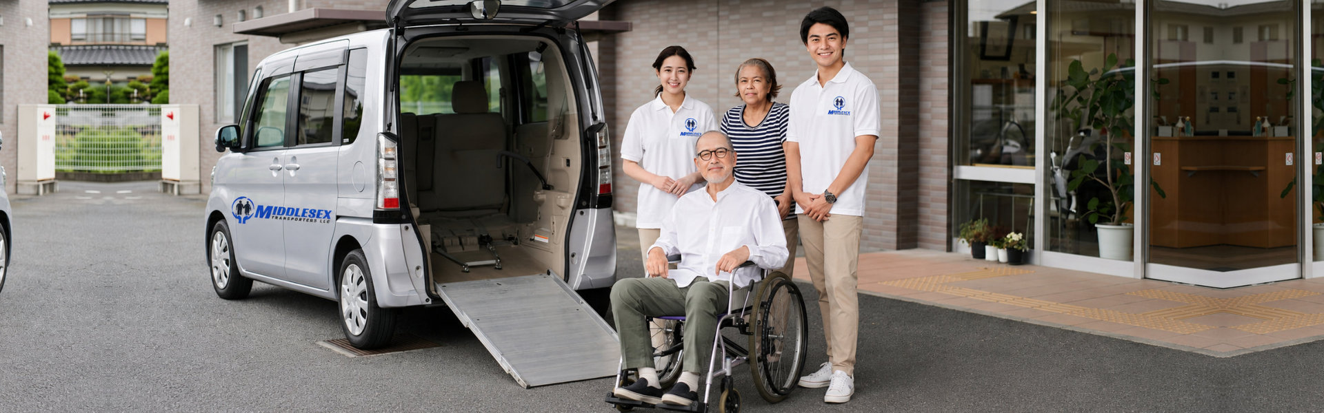 A group of caregivers assist a senior in a striped shirt and a man in a wheelchair near a van. Two young caregivers in blue polo shirts stand behind them. A silver van has a ramp extended. The location appears to be a hospital or healthcare facility. The overall mood is light-hearted, conveying a sense of support and care. This image would be ideal for projects related to assisted living, home healthcare, or medical transportation services. It could also be used to illustrate themes of compassion, community, and aging with dignity.