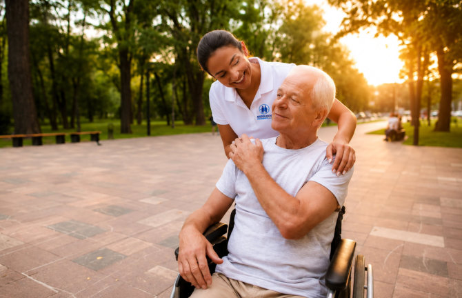The old man who sits in a wheelchair holds the nurse by the hand, who laid her hand on his shoulder 