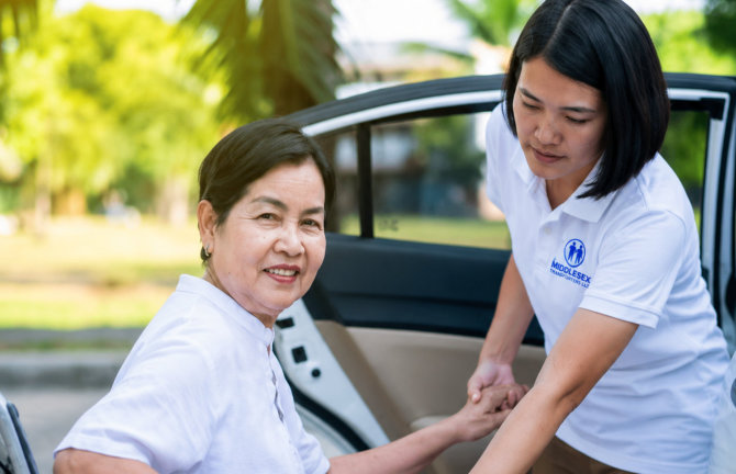 Caregiver helping senior handicapped asian woman from wheelchair get into car 
